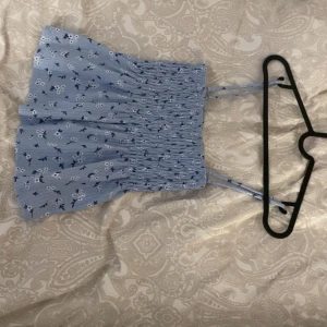 Blå blommig croppad linnetopp från Gina Tricot - Söt ljusblå croppad linnetopp från Gina Tricot med små vita och mörkblå blommor. Toppen har smock över hela livet och tunna axelband. Perfekt för varma dagar!