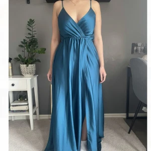 Blå långklänning med slits och omlott - Säljer en elegant blå långklänning.  Klänningen har en markerad midja och en hög slits framtill som ger en snygg siluett. Aldrig använd! Passar både S och M