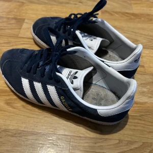 Adidas Gazelle sneakers i marinblå mocka - Säljer ett par klassiska Adidas Gazelle sneakers i marinblå mocka med vita detaljer och de ikoniska tre ränderna på sidorna. Skorna har snörning och vit sula, samt Adidas-logga på plösen och hälen. Storlek:34 men stora i storleken passar 35 