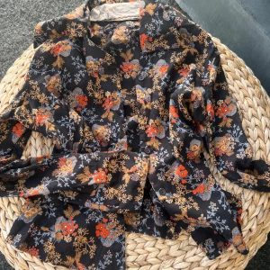 Svart blommig skjorta från BronZella - Säljer en svart skjorta från BronZella med färgglatt blommigt mönster i orange, gult och grått. Skjortan har långa ärmar, knappar framtill och en fin volang längst ned. Perfekt för dig som gillar unika och färgstarka plagg! På lappen står st 36, men skulle snarare säga st 32.