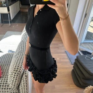 Svart kort klänning med volangkjol från Ralph Lauren - Supersnygg svart klänning från Ralph Lauren med krage, knappar framtill och korta ärmar. Tight passform med markerad midja och bälte med silverdetalj. Kjolen har flera lager volanger, jag är 180 lång som jämförelse hur den sitter