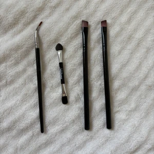Fyra sminkborstar och applikator - Säljer ett set med fyra sminkverktyg: två platta borstar med svarta skaft, en vinklad tunn borste och en dubbeländad ögonskuggsapplikator med svart och silverfärgat handtag. Perfekta för detaljerad ögonmakeup och skuggning.
