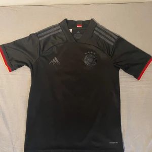Svart Tyskland fotbollströja Adidas - Snygg svart Tyskland fotbollströja från Adidas med landslagsemblem och fyra stjärnor på bröstet. Tröjan har gråa ränder på axlarna, korta ärmar med röd och gul detalj vid ärmslut samt tillverkad i lätt och ventilerande Aeroready-material.