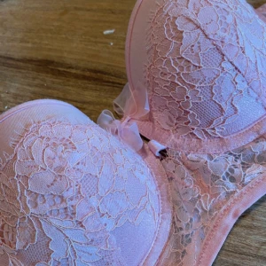 Rosa spetsig bh - Söt ljusrosa bh med vadderade kupor och vacker spets över hela framsidan. Toppen har en liten rosett framtill och transparent mesh på sidorna. Perfekt för dig som gillar romantiska detaljer och mjuka pasteller.