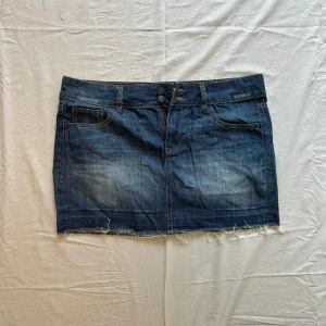 Mörkblå jeanskjol med fransig kant -  Rakt över midjan: 44 cm , längd: 35 cm Snygg mörkblå jeanskjol med klassiska fickor fram och bak, dubbla knappar i midjan och fransig nederkant