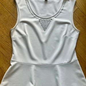 Vit topp med strass från MQ Edition - Vit topp från MQ Edition med rund halsringning dekorerad med strass i ett triangulärt mönster. Toppen har en figurnära passform och är sydd i ett mjukt, stretchigt material. Perfekt för dig som gillar stilrena detaljer och lite extra bling.