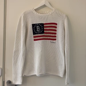 Vit stickad tröja Bondelid S - Vit stickad tröja från Bondelid i storlek S. Tröjan har lång ärm och ett broderat amerikanskt flaggmotiv med Bondelid-logga på bröstet. Klassisk rund halsringning och ribbade muddar.