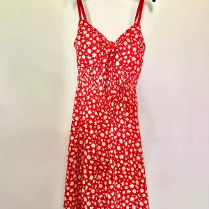 Röd blommig klänning  - Superfin röd klänning med vita blommor, smala justerbara axelband och söt knytning framtill samt uttagbara inlägg. Midja i resor ger en skön passform. Materialet är tunt och svalt men inte genomskinligt. Storleken är unisex men passar mig som är strl. M/38 och 172 cm lång men passar även strl. S/36 och L/40 bla.