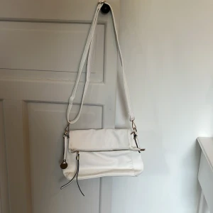 Vit axelväska i skinn med dragkedja - Snygg vit axelväska i skinn med justerbart axelband och guldfärgade detaljer. Väskan har dragkedja framtill, dekorativa band och en liten ficka med dragkedja på baksidan. Invändigt finns ett mörkblått foder och extra fack för småsaker. Bara att skriva vid frågor. Pris går att förhandla!