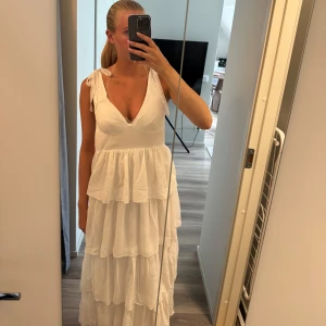 Vit långklänning med volanger - Superfin vit långklänning med flera lager volanger och v-ringning. Axelbanden knyts med rosett och klänningen har en luftig, flowy känsla. Perfekt för sommarens alla tillfällen och ger en romantisk vibe.