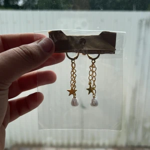 Guldiga örhängen med stjärnor och pärlor - Snygga guldiga örhängen med kedjor, små stjärnhängen och droppformade vita pärlor. Perfekta för dig som vill ha något unikt och trendigt med lite extra detalj. Lätta att matcha med olika stilar och outfits.