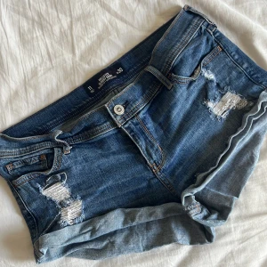 Blå slitna jeansshorts Hollister - Snygga blå jeansshorts från Hollister med låg midja och avslappnad passform. Shortsen har slitna detaljer framtill och uppvikta benslut för en chill vibe. Klassiska fickor både fram och bak, perfekt till sommaren.