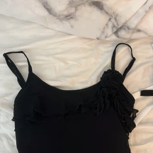 Svart linnetopp med volang och blomdetalj - Supersnygg svart linnetopp med tunna axelband, volang längs bröstet och en stor tygblomma på ena axeln. Toppen har asymmetrisk nederkant och är gjord i ett lätt, skirt material som ger en cool och luftig känsla.