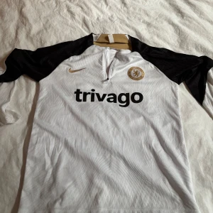 Chelsea långärmad tränings-topp Nike - Chelsea FC långärmad tränings-tröja från Nike i vitt med svarta ärmar och guldiga detaljer. Tröjan har halv dragkedja, broderad Nike-logga och Chelsea-märke på bröstet. Materialet är lätt och andas, perfekt för träning. Trivago-tryck på framsidan. Pasformen är lite konstig vet ej om den är från Nike