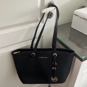 Svart axelväska  - Snygg svart axelväska liknande Michael Kors med guldfärgade detaljer och MK-hänge. Väskan har dragkedja upptill och långa axelband, perfekt för att bära allt du behöver. Stilren design som passar till många outfits. Två fack inuti och en yttre sidofack.