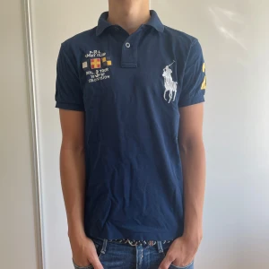 Piké från Polo Ralph Lauren - Säljer en marinblå pikétröja från Polo Ralph Lauren med broderade detaljer. Tröjan har ett stort polo märke, siffror vid axeln och även ett Yacht club märke. Denna piké är riktigt stilren och perfekt nu på sommaren! Tveka inte på att skriva om du har frågor! 🙌Priset är inte hugget i sten 🪨 