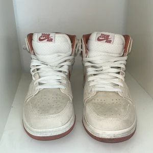 Nike Air Jordan 1 High Zoom Air beige/röd - Nike Air Jordan 1 High Zoom Air i beige mocka med röda detaljer och swoosh. Skorna har perforerad tå, vit snörning och röd logga på plösen. Insidan och yttersulan är röd, vilket ger en snygg kontrast till den ljusa ovandelen. Perfekta för dig som gillar klassiska sneakers med unik färgkombo.