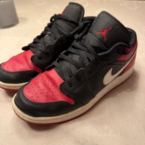 Nike Air Jordan 1 Low Bred - Säljer ett par Nike Air Jordan 1 Low i den klassiska Bred-färgställningen. Skorna har svart och rött skinn, vit swoosh och röd yttersula. Ikonisk Air Jordan-logga på hälen och röd Jumpman på plösen. Perfekta för dig som gillar streetwear och sneakers med retrovibe.