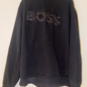 Svart sweatshirt från BOSS - Svart sweatshirt från BOSS med stor, glansig BOSS-logga framtill. Tröjan har en klassisk passform med långa ärmar och rund hals. Perfekt för dig som gillar stilrena och snygga plagg med tydlig logga. Snabb affär kan diskuteras 