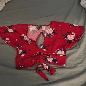 Röd blommig omlottopp från H&M - Superfin röd omlottopp från H&M med blommigt mönster i vitt och mörkblått. Toppen har korta vida ärmar, v-ringning och knyt framtill. Perfekt för dig som vill sticka ut med färg och mönster. Snygg till både jeans och kjol!