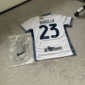 Inter Milan Barella 23 Nike fotbollströja - Inter Milan bortatröja med Barella 23 på ryggen. Tröjan är vit med marinblå detaljer, korta ärmar och klassisk krage. Officiella klubbmärken, sponsortryck och Nike-logga framtill. Tillverkad i lätt och ventilerande material, perfekt för match eller träning.