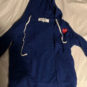 Blå hoodie från Comme des Garçons Play - Blå hoodie från Comme des Garçons Play med dragkedja och vit snörning i huvan. Ikoniskt rött hjärta med ögon på bröstet. Mjuk bomullskänsla och klassisk passform, perfekt för en chill och stilren look., Storlek S men sitter som XS