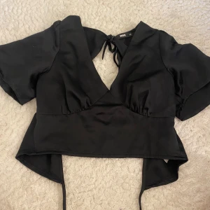 Svart omlottopp från Bik Bok - Svart omlottopp från Bik Bok med korta ärmar och djup v-ringning. Toppen har knytband i ryggen och är tillverkad i ett mjukt, glansigt material som ger en lyxig känsla. Perfekt för dig som gillar trendiga och stilrena plagg.