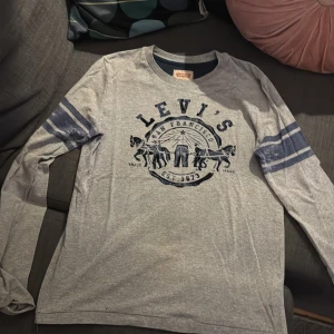 Grå långärmad Levi's tröja - Snygg grå långärmad tröja från Levi's med blå tryck framtill och coola blå ränder på ärmarna. Klassisk rund halsringning och avslappnad passform. Perfekt för dig som gillar streetstyle och vill ha något bekvämt men ändå stilrent.