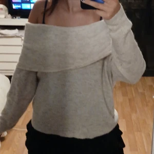 Beige offshoulder stickad tröja - Supermysig beige stickad tröja med offshoulder-design och långa ärmar. Tröjan har en bred krage som vilar snyggt över axlarna och ger en chill vibe. Perfekt för lager-på-lager under hösten eller vintern. Passar dig som gillar en lite loose fit.