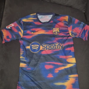 FC Barcelona Lamine Yamal matchtröja - Säljer en FC Barcelona matchtröja med Lamine Yamal och nummer 19 på ryggen. Tröjan har ett färgstarkt mönster i blått, rött, gult och orange, med klubbmärke, Nike-logga och Spotify som sponsor framtill. Tillverkad i lätt och ventilerande material, perfekt för fotbollsträning.