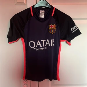 FC Barcelona Messi 10 matchtröja - Snygg FC Barcelona fotbollströja med Messi 10-tryck på ryggen och Qatar Airways-logga på bröstet. Tröjan är mörkblå med svarta ärmar och röda detaljer längs sidorna och ärmsluten. Broderat klubbmärke på bröstet. Tillverkad i lätt och ventilerande material.