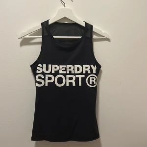 Svart sportigt linne  - Svart linnetopp från Superdry med stort vitt tryck framtill där det står 'SUPERDRY SPORT'. Modellen är ärmlös med brottarrygg och tillverkad i ett stretchigt syntetmaterial som andas, perfekt för träning och aktiva dagar. Linnet har en inbyggd ”sport bh” som håller brösten på plats. 