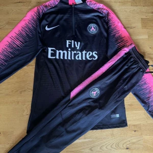 PSG Nike tracksuit svart/rosa S - Säljer ett par svarta PSG träningsbyxor från Nike med rosa detaljer längs sidorna och broderad klubbmärke. Byxorna är i sportig Dri-FIT polyester, har elastisk midja med snörning och smal passform. Perfekta för fotbollsträning eller chill.