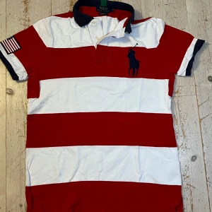 Randig pikétröja från Polo Ralph Lauren - Snygg pikétröja från Polo Ralph Lauren i rött och vitt med breda ränder. Tröjan har klassisk krage i mörkblått, broderad logga på bröstet och amerikansk flagga på ärmen. Tillverkad i bomull och har korta ärmar. Nypris 2100