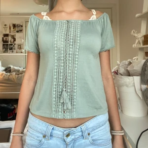 Ljusgrön offshoulder blus med spets - Superfin ljusgrön offshoulder blus med korta ärmar och spetsdetaljer framtill. Blusen har dekorativ snörning med tofsar och små bollar vid ärmsluten. Perfekt för en chill och somrig look.