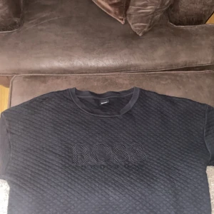 Svart quiltad tröja från Hugo Boss - Säljer en svart quiltad tröja från Hugo Boss med stort broderat BOSS-logo framtill. Tröjan har rund halsringning och är tillverkad i mjukt material som känns skönt mot huden. Perfekt för dig som gillar stilrena och exklusiva plagg med tydlig logga.