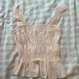 Ljusrosa smockad linnetopp H&M XS - Supersöt ljusrosa linnetopp från H&M i storlek XS. Toppen har breda axelband, smockad resår över hela livet och en volangkant nertill. Perfekt för varma dagar och ger en romantisk vibe till din outfit.