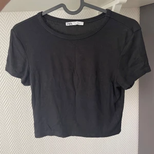 Svart croppad topp från Zara - Enkel och stilren svart croppad topp från Zara. T-shirttyp med rund hals och korta ärmar, perfekt att matcha med höga jeans eller kjol. Mjuk bomullskänsla och klassisk passform som funkar till många olika looks.