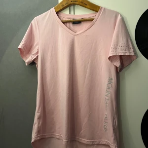 Ljusrosa t-shirt från Mountain Horse - Snygg ljusrosa t-shirt från Mountain Horse med v-ringning och korta ärmar med perforerade detaljer. Tillverkad i mjuk bomull, perfekt för dig som gillar en avslappnad och sportig stil. Diskret logga längs sidan ger en clean look. Notera liten fläck på framsidan 