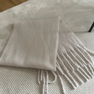 Beige fluffig halsduk med fransar - Supermjuk och fluffig beige halsduk med långa fransar i kanterna. Perfekt att svepa runt halsen för extra värme och stil. Enkel design som passar till det mesta och ger en clean look.