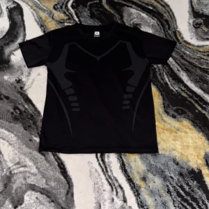 Svart sportig t-shirt i funktionsmaterial - Svart t-shirt i funktionsmaterial med coolt grafiskt mönster framtill. Perfekt för träning eller när du vill ha en sportig look. Klassisk rund hals och korta ärmar, tillverkad i lätt och snabbtorkande syntetmaterial.