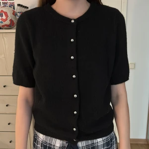 Svart kofta med pärlknappar från H&M - Svart stickad kofta från H&M i storlek S. Kortärmad modell med rund halsringning och snygga pärlknappar framtill. Mjuk och skön, perfekt att slänga över en topp eller klänning. Enkel och stilren design som funkar till det mesta. Använd ett fåtal gånger.