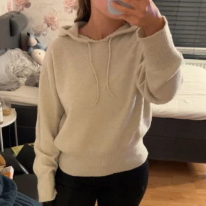 Beige stickad hoodie med huva - Mysig beige stickad hoodie med huva och snörning. Tröjan har långa ärmar och ribbade muddar vid ärmslut och nederkant. Perfekt för dig som gillar en avslappnad och stilren look.