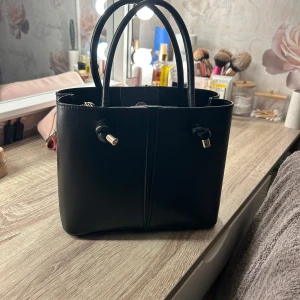 Svart handväska med dragkedja - Stilren svart handväska i slätt skinnliknande material med två handtag och snygga metall-detaljer. Väskan har en praktisk innerficka med dragkedja och knäppning upptill. Perfekt storlek för det viktigaste och passar till många outfits.