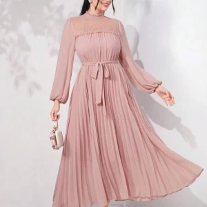 Ljusrosa långklänning med spets - Superfin ljusrosa långklänning med plisserad kjol, transparent spets upptill och högt halsad krage. Klänningen har långa, luftiga ärmar och ett knytband i midjan för extra snygg passform. Perfekt för dig som vill ha en romantisk och elegant look.