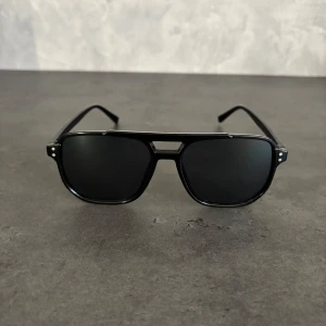 Svarta pilotbågar solglasögon - Snygga svarta solglasögon med pilotbågar och mörka linser. Ramen är i blank svart plast med coola silvriga nitar vid hörnen. Klassisk och stilren design som ger en edgy vibe till din look.