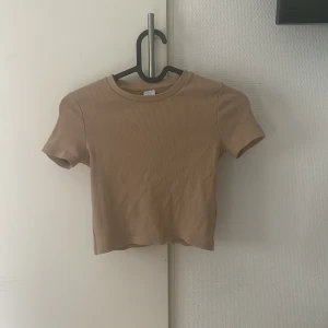 Beige ribbad croppad topp Lindex - Säljer en beige ribbad croppad topp från Lindex. T-shirten har korta ärmar, rund halsringning och är tillverkad i mjuk bomull. Perfekt för en chill och enkel stil.