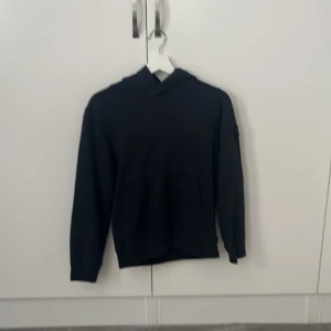 Svart hoodie från Stone Island Junior - Svart hoodie från Stone Island Junior med klassisk logga på ärmen. Tröjan har huva, långa ärmar och en enkel, clean design. Perfekt för dig som gillar streetwear och vill ha något stilrent men ändå med tydlig attityd.