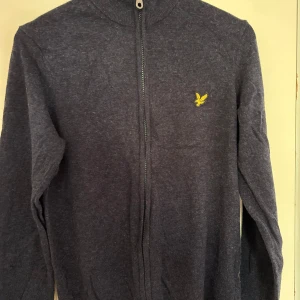 Mörkgrå kofta från Lyle & Scott - Mörkgrå kofta från Lyle & Scott med hel dragkedja och klassisk gul logga på bröstet. Tröjan är stickad och har lång ärm samt ribbade muddar vid hals och ärmslut. Perfekt för lager-på-lager och snygg till jeans eller chinos.