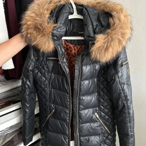 Svart Rockandblue dunjacka med päls - Snygg svart dunjacka från Rockandblue med stor fluffig räv- päls på huvan och guldfärgade dragkedjor. Jackan har quiltade detaljer, innerfoder med leopardmönster och flera praktiska fickor. Perfekt för kalla dagar när du vill vara både varm och stilren.